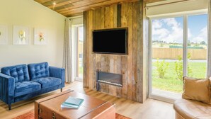 A living room with a blue sofa, a wooden entertainment unit with a TV, and a fireplace. - New in 2022 Luxurious and contemporary lodge with stunning countryside views. (Bretby, Derbyshire)