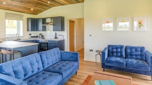 A modern living room with a blue sofa, a kitchen with dark cabinets, and a wooden ceiling. - New in 2022 Luxurious and contemporary lodge with stunning countryside views. (Bretby, Derbyshire)