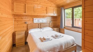 A wooden cabin bedroom with a bed, nightstands, a window, and a wall-mounted clock. - Comfortable and spacious lodge with two doubles and one with bunks. (Glenluce, Newton Stewart)