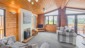 Een knus cabin interieur met een open haard, bank, televisie en grote ramen met uitzicht op de omgeving.