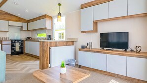 A modern kitchen with a built-in oven, a dining area with a wooden table, and a TV on a wooden stand. - Signature 2 Bed Caravan (Ormside, Appleby-in-Westmorland)