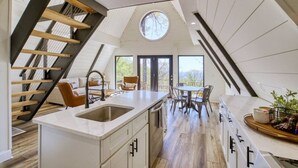 A modern kitchen with a central island, a dining area with a round table, and a staircase with wooden beams.