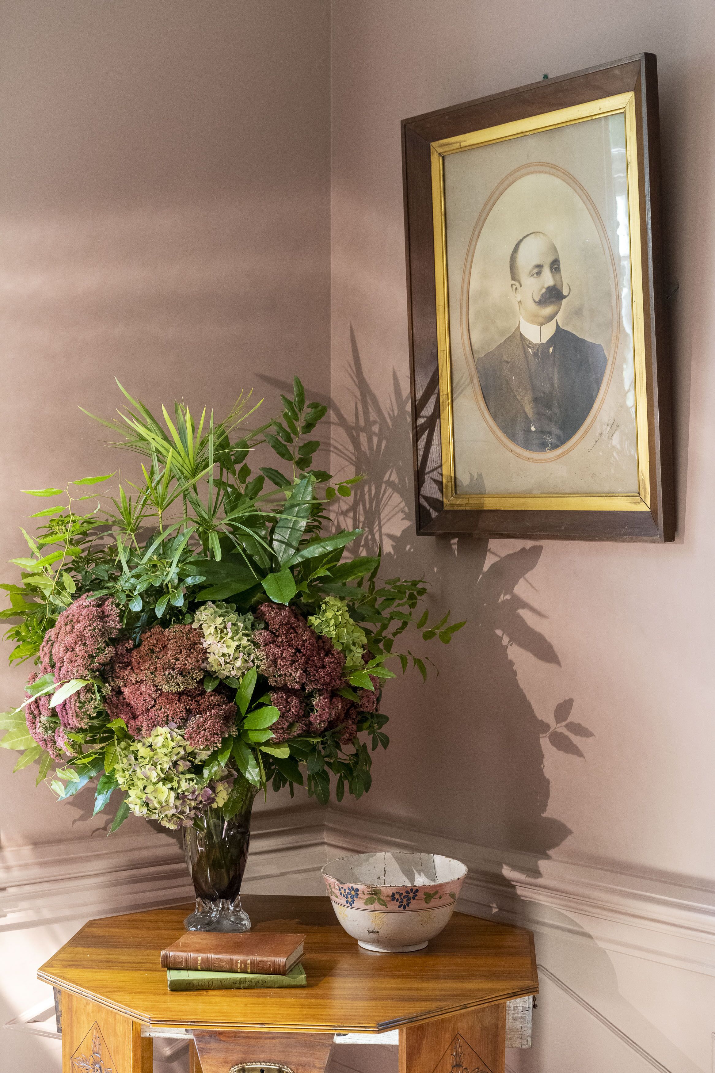 A wooden table with a vase of flowers, a bowl, and books. A framed portrait hangs on the wall.