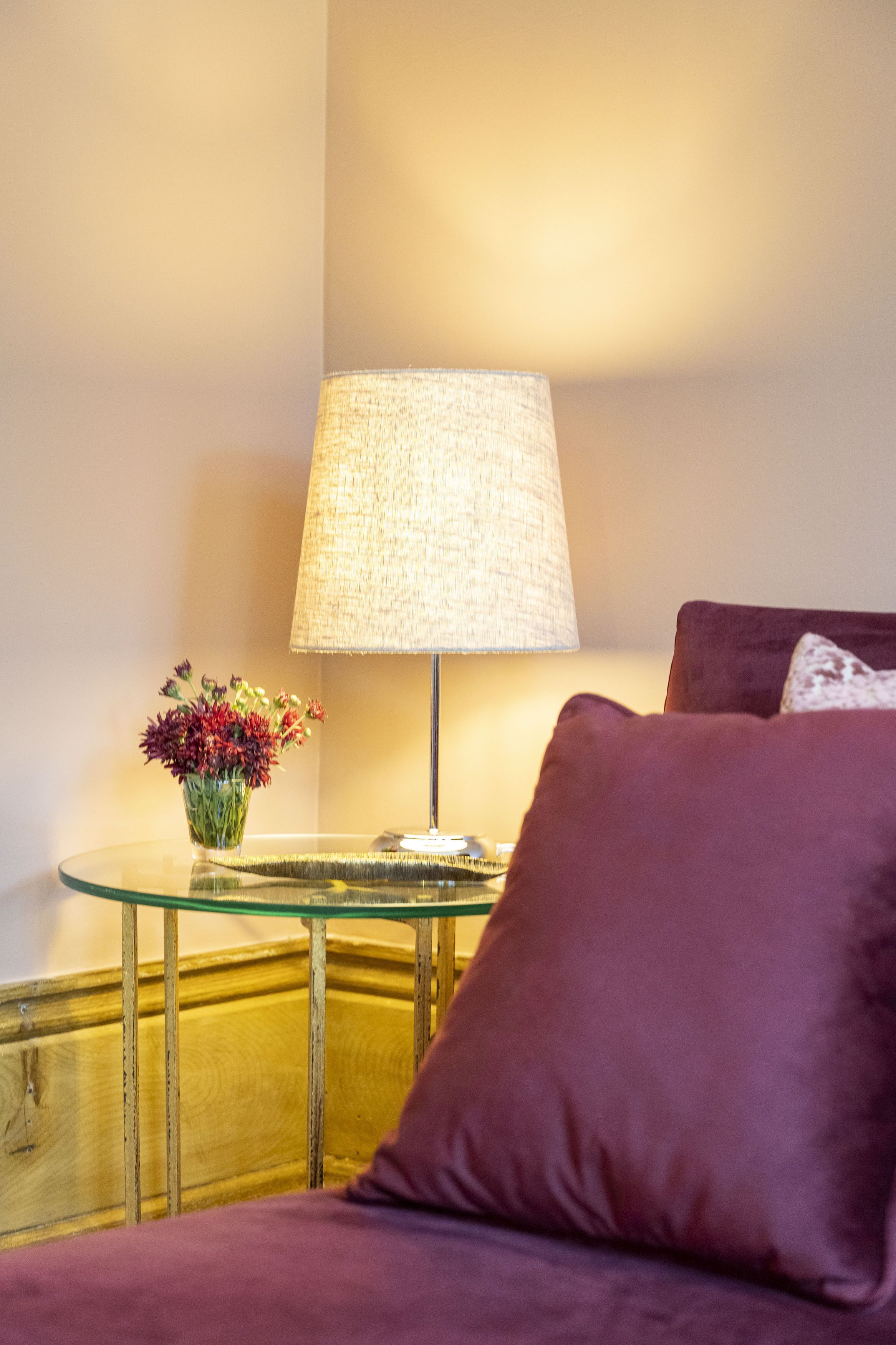 Une pièce avec une table en verre, une lampe, un vase de fleurs et un coussin violet.