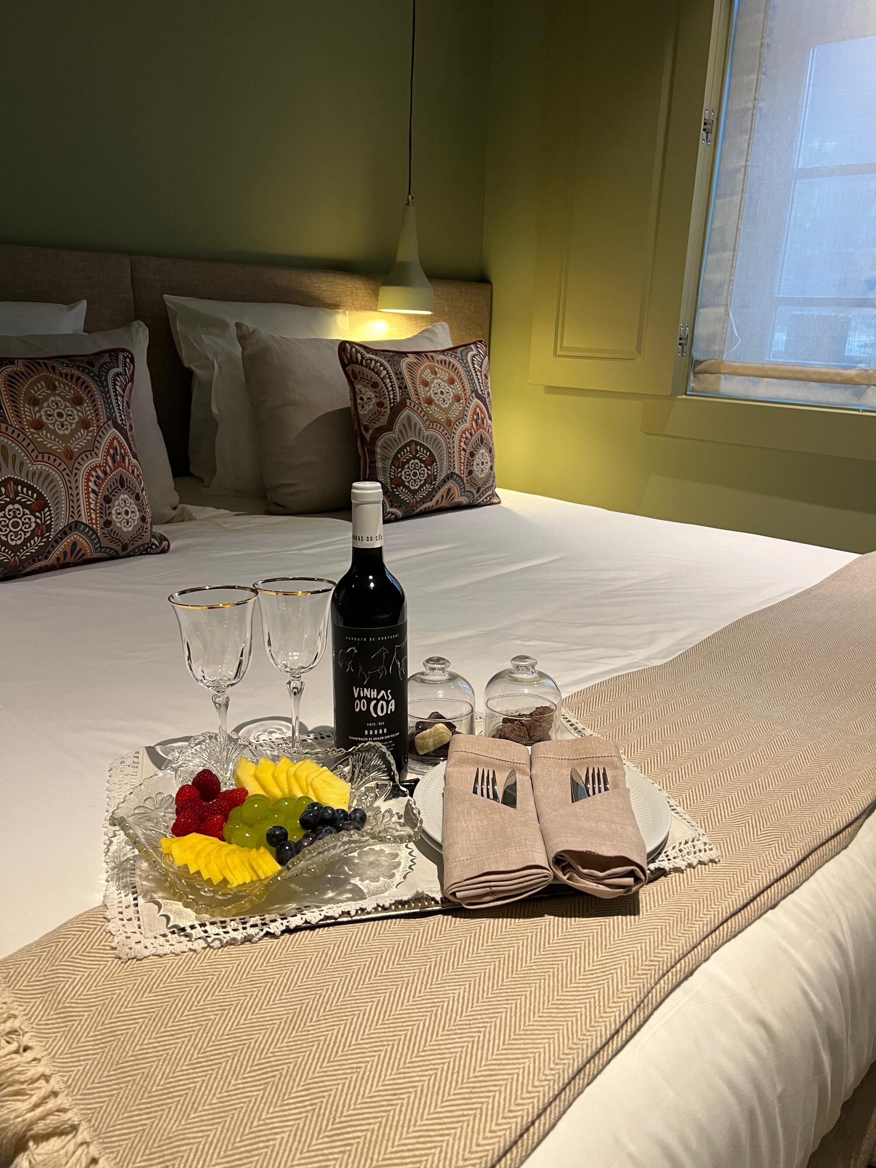 Un lit bien fait, avec une bouteille de vin, des verres et une assiette de fruits.