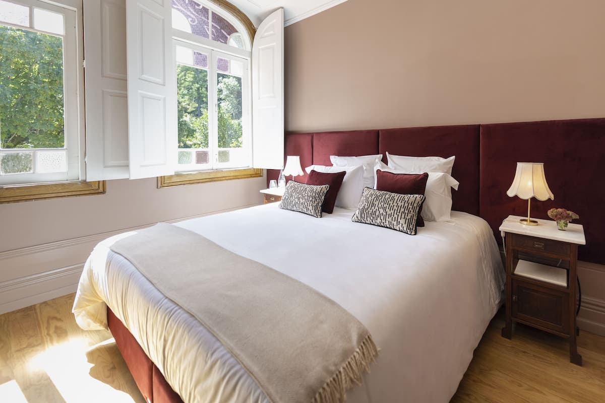 A large bed with a white and beige bedspread, two patterned pillows, a red headboard, and a nightstand with a lamp and vase.