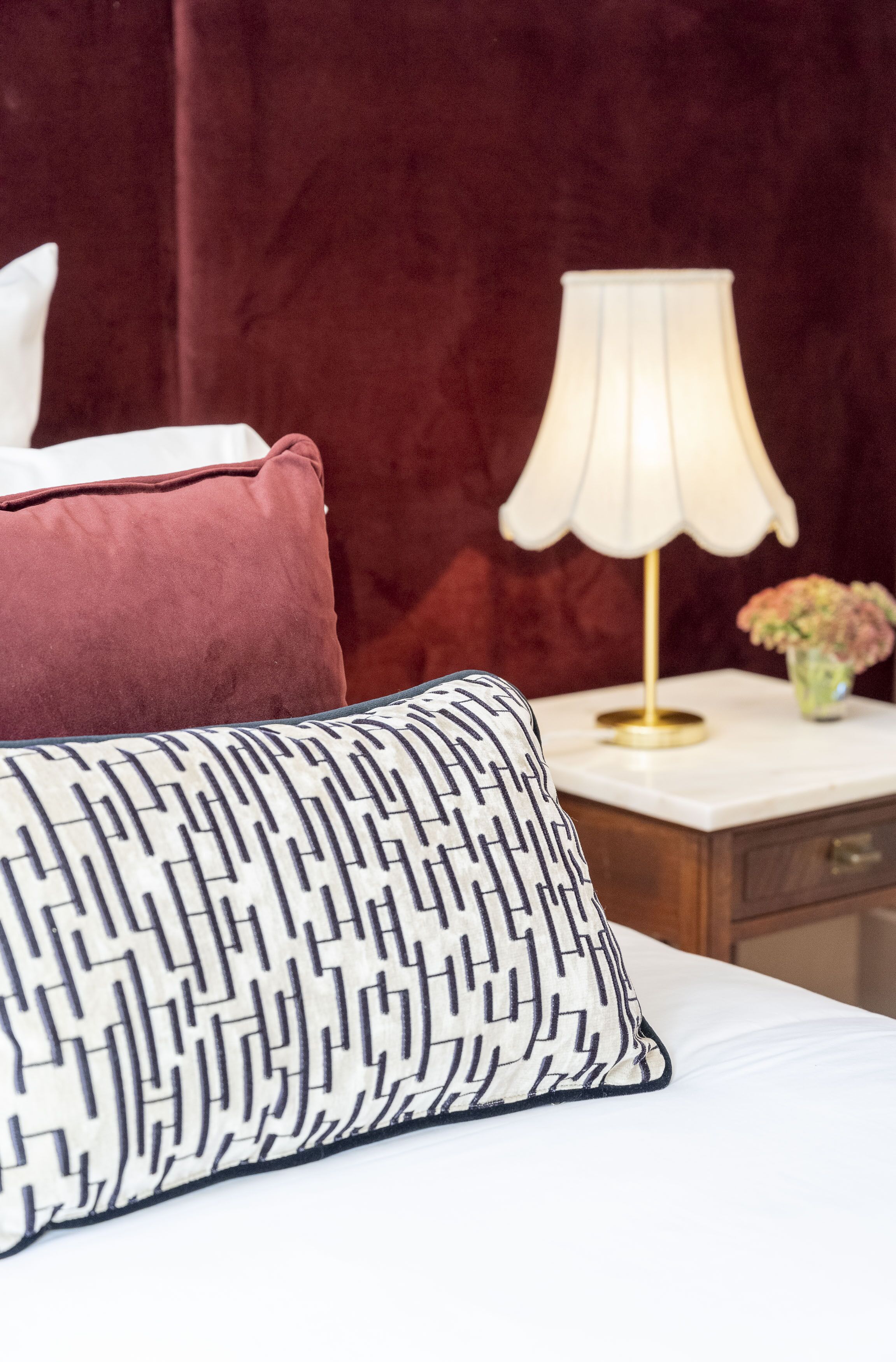 A bed with a patterned pillow and a red cushion, a bedside lamp, and a vase with flowers on a nightstand.