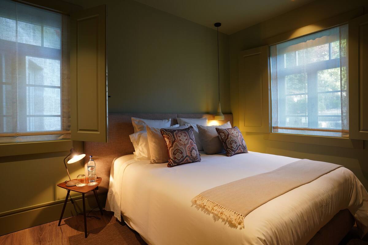 A well-lit bedroom with a large bed, multiple pillows, a bedside table with a lamp, and a window providing natural light.