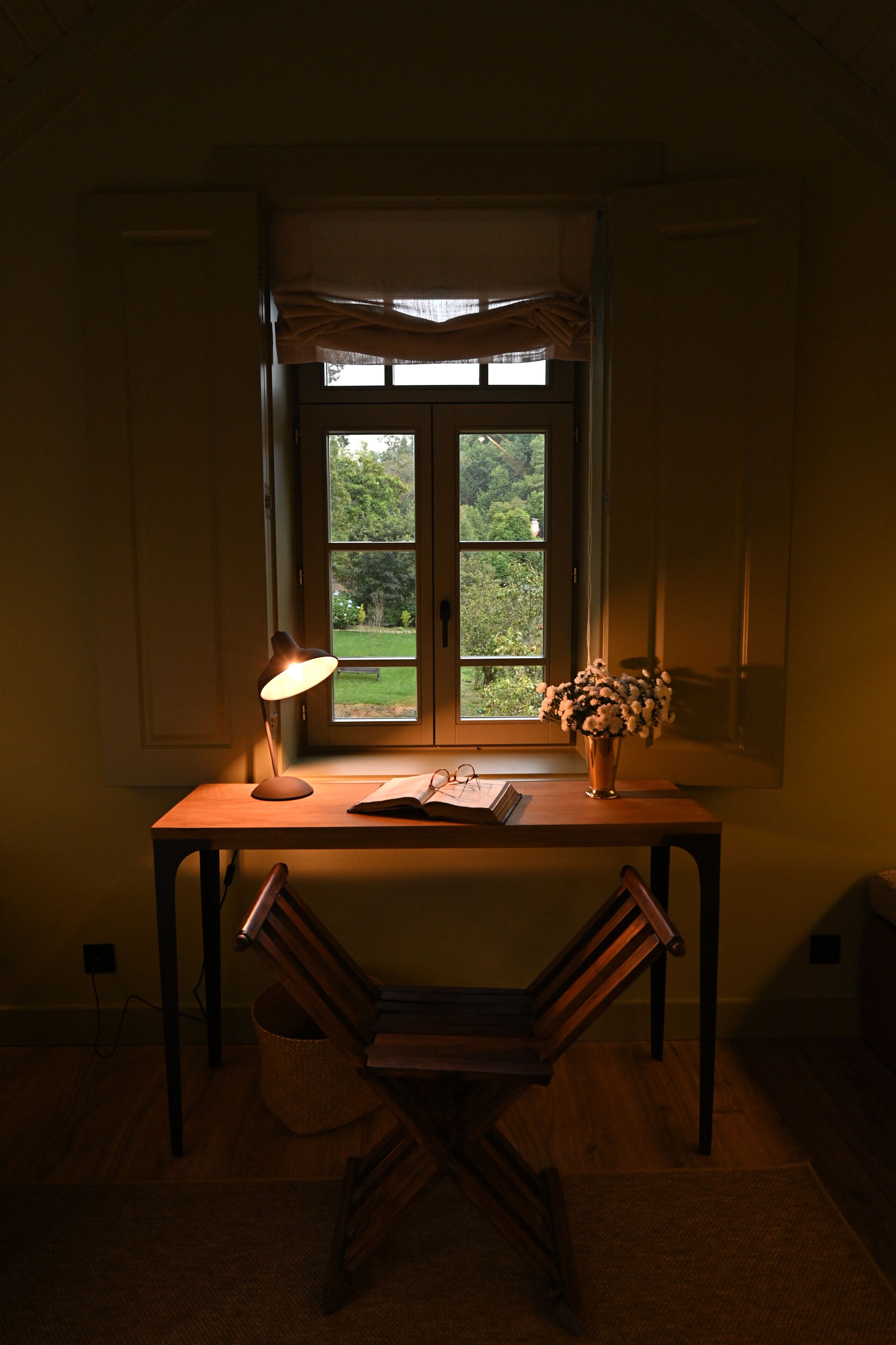 A room with a desk, a lamp, a chair, a window with a view of greenery, and a vase with flowers.