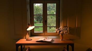 A room with a desk, a lamp, a chair, a window with a view of greenery, and a vase with flowers.
