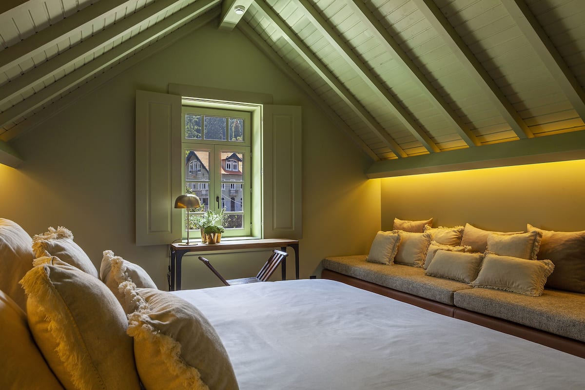 A bedroom with a large bed, a window with a view of houses, a small table with a lamp, and a sofa with cushions.