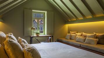 Un dormitorio con una cama grande, una ventana con vistas a casas, una mesita de noche con lámpara y un sofá con cojines.