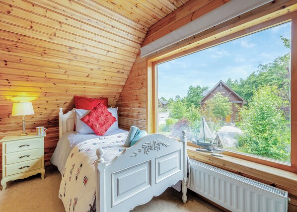 Une chambre avec un plafond en pente en bois, un lit avec une tête de lit blanche, une table de chevet avec une lampe et une fenêtre donnant sur une maison et des arbres.