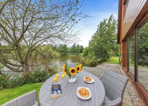 Una mesa redonda con platos de comida, un libro y un jarrón con girasoles, con vistas a un cuerpo de agua y follaje.
