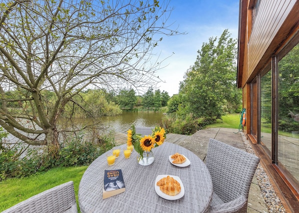 Une table ronde, sur laquelle on peut voir des assiettes remplies de nourriture, un livre et un vase avec des tournesols, le tout donnant sur un plan d’eau et une végétation luxuriante.