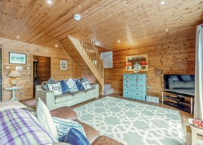 A cozy cabin interior with a wooden ceiling, a sofa with patterned cushions, a television, and a staircase.