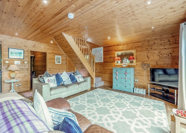 A cozy cabin interior with a wooden ceiling, a sofa with patterned cushions, a television, and a staircase. - Ground floor: Spacious open plan lounge, kitchen, dining area with log burner. (Anglesey)