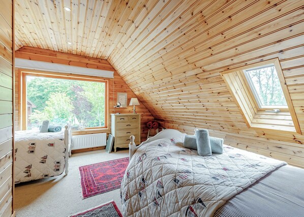 Une chambre avec un plafond en pente, un lit recouvert d’une courtepointe matelassée, une commode, une table de chevet et une fenêtre donnant sur les arbres.