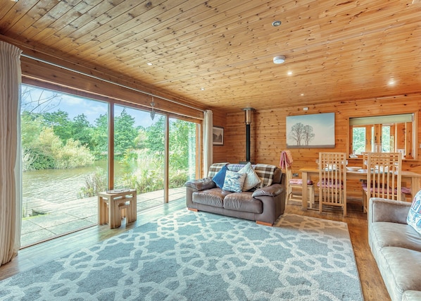 A wooden cabin interior with a sofa, dining table, chairs, and large windows overlooking a natural landscape. - Ground floor: Spacious open plan lounge, kitchen, dining area with log burner. (Anglesey)