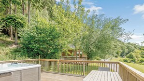A wooden deck with a hot tub, a table, and chairs, surrounded by lush greenery and trees.