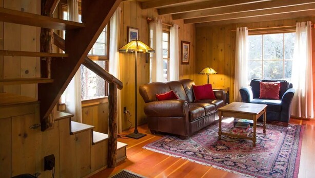 A cozy cabin interior with a wooden staircase, leather sofas, and a large window. - Twin Rock Tree House - Vintage Cabin (Idyllwild-Pine Cove)