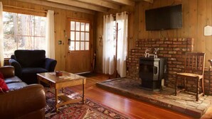 A cozy cabin interior with a wooden floor, a fireplace, a television mounted on the wall, and a large window with curtains. - Twin Rock Tree House - Vintage Cabin (Idyllwild-Pine Cove)