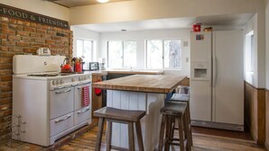 A kitchen with a brick wall, a stove, a refrigerator, and a wooden island with stools. - Twin Rock Tree House - Vintage Cabin (Idyllwild-Pine Cove)