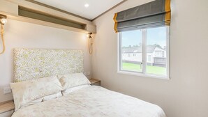 A bedroom with a bed, a headboard with a floral pattern, a window with a view of houses, and a bedside table with a lamp.