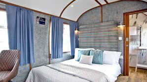 A bedroom with a bed, a chair, a window with curtains, a headboard, and a bathroom with a mirror and towel rack.