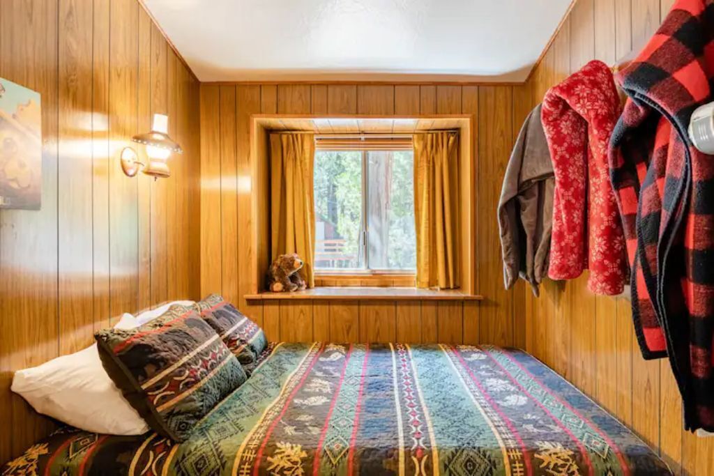 Un cuarto acogedor con paredes de madera, una ventana con cortinas, una cama con ropa de cama estampada y un osito de peluche en el alféizar.