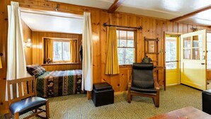 Una habitación acogedora con paredes de madera, una cama con ropa de cama estampada, un sillón de cuero negro, una mesita pequeña y una puerta amarilla.