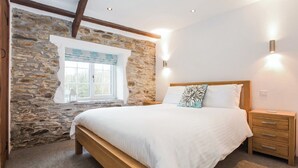 A bedroom with a stone wall, a wooden bed frame, a nightstand, and a window with a view. - 2 bedroom accommodation in Watergate, Looe (Watergate, Looe)