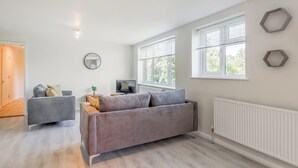 Ein modernes Wohnzimmer mit einem grauen Sofa, einem Fernseher und zwei Fenstern mit Jalousien.