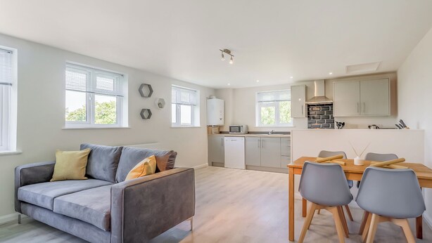 Eine moderne Wohnung mit grauem Sofa, Esstisch und Stühlen, einer Küche mit weißen Schränken und großen Fenstern.