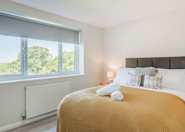 A bedroom with a large window, a bed with a beige blanket, a bedside lamp, and a radiator.