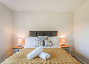 Un lit bien fait, recouvert d’une couvre-lit à rayures, avec deux oreillers et deux serviettes enroulées. Deux lampes de chevet sont posées sur des tables de nuit en bois.