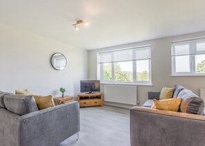 Un salon moderne avec des canapés gris, un téléviseur, une petite table d’appoint en bois et une grande fenêtre avec des stores.