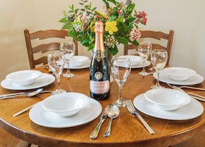 A round wooden dining table set for a meal with white plates, silverware, and wine glasses, centered around a bottle of Camelfley Rose Brut.