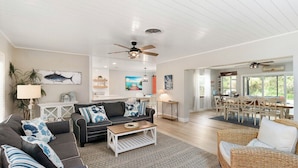 A spacious living room with a dark grey sofa, a white coffee table, and a wicker chair. There is a ceiling fan, a dining area with a table and chairs, and a view of the outdoors through large windows. - Heated pool on the water - under 10 mins to beaches! (Sarasota)