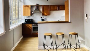 Une cuisine moderne avec des armoires en bois, un four encastré, un four à micro-ondes et une cuisinière à gaz. On y trouve trois tabourets en bois et un luminaire suspendu.