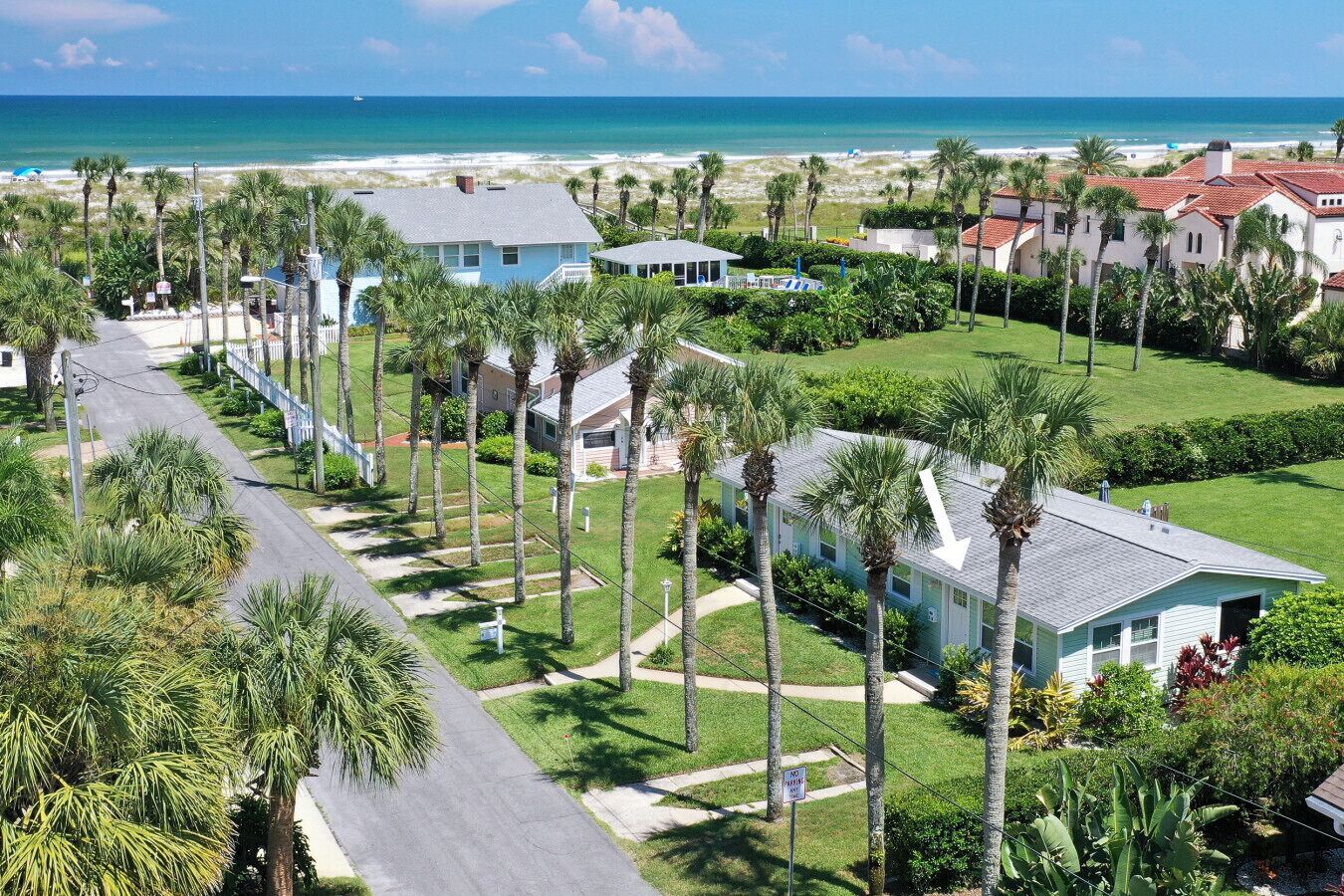 A coastal residential area with a road lined by palm trees and houses, leading to a beach.