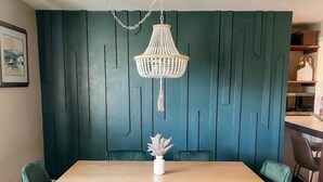 Una zona de menjador amb una taula de fusta, cadires tapissades de verd, un rameller blanc amb flors i un candelabrum blanc.