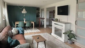 Un saló amb un sofà de pell marró, una taula de centre de fusta, una zona de menjador amb taula i cadires, una llar de foc blanca i un televisor fixat a la paret.