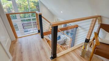 A spacious loft bedroom with a wooden floor, a wooden staircase, a large window, and a view of the outdoors.