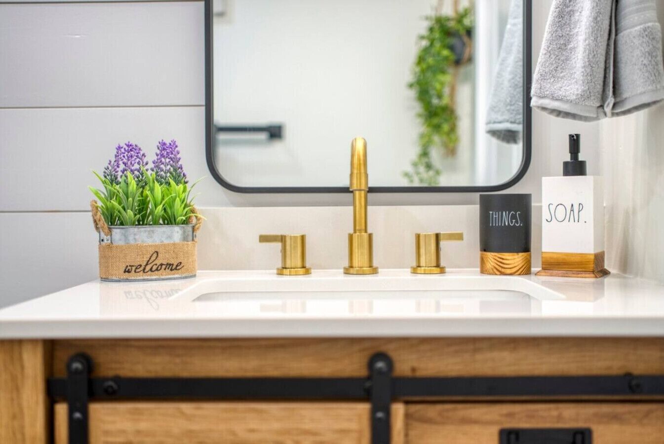 A bathroom vanity with a white countertop, a gold faucet, a soap dispenser, and a welcome sign with purple flowers.