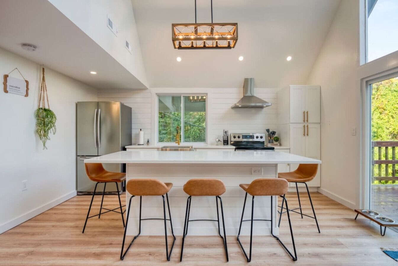Een moderne keuken met een centraal eiland, barkrukken, een roestvrijstalen koelkast en een groot raam met uitzicht op groen.
