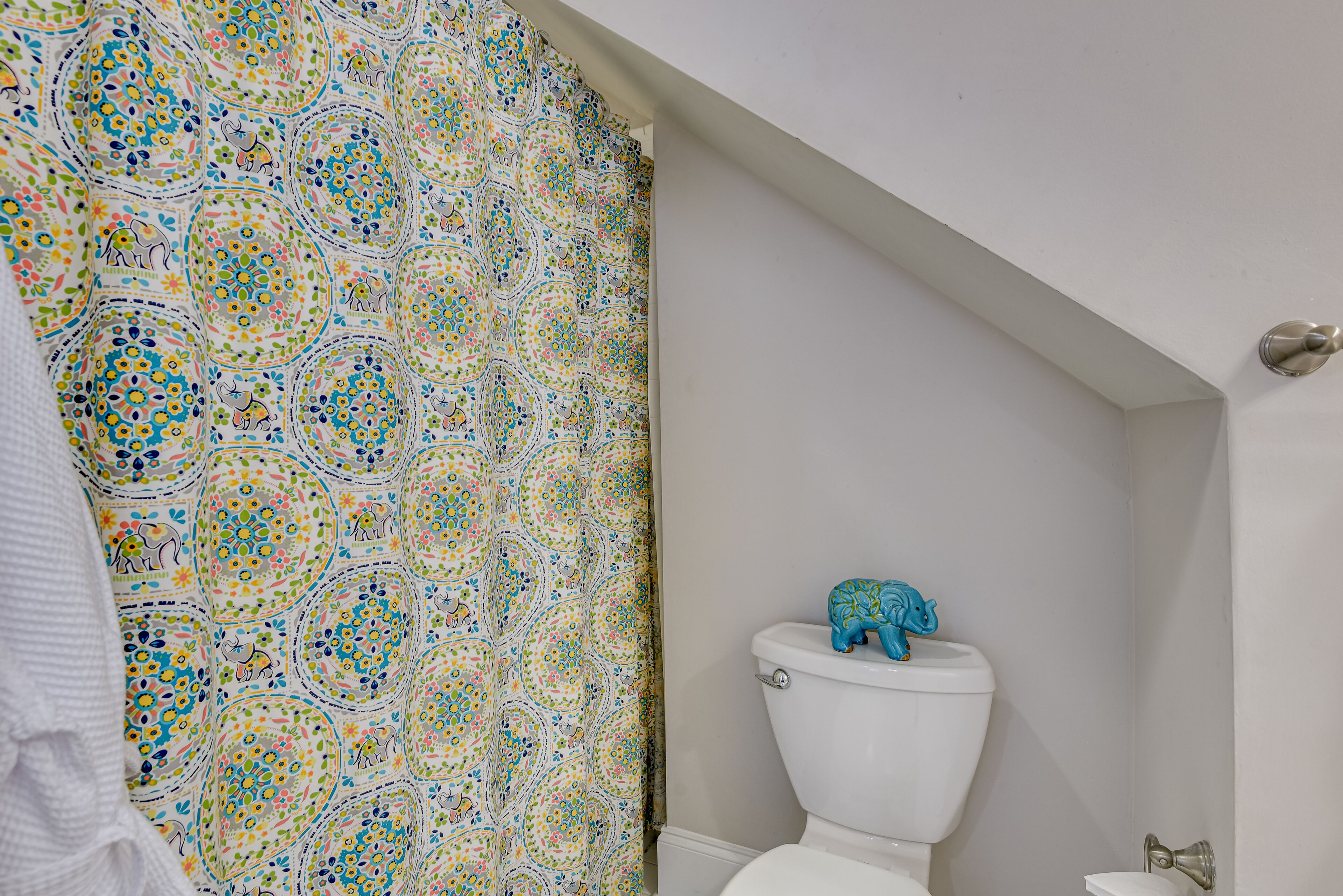 A bathroom with a patterned shower curtain, a white toilet, and a blue elephant decoration on the tank.
