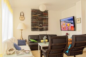 Una sala de estar con un sofá, dos sillones, una mesa de centro, una lámpara, un tapiz en la pared, un televisor y un cuadro enmarcado.
