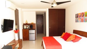 Habitación de hotel con cama, escritorio, televisión, ventilador de techo, refrigerador, microondas, un florero con flores y una puerta que da a otra habitación.