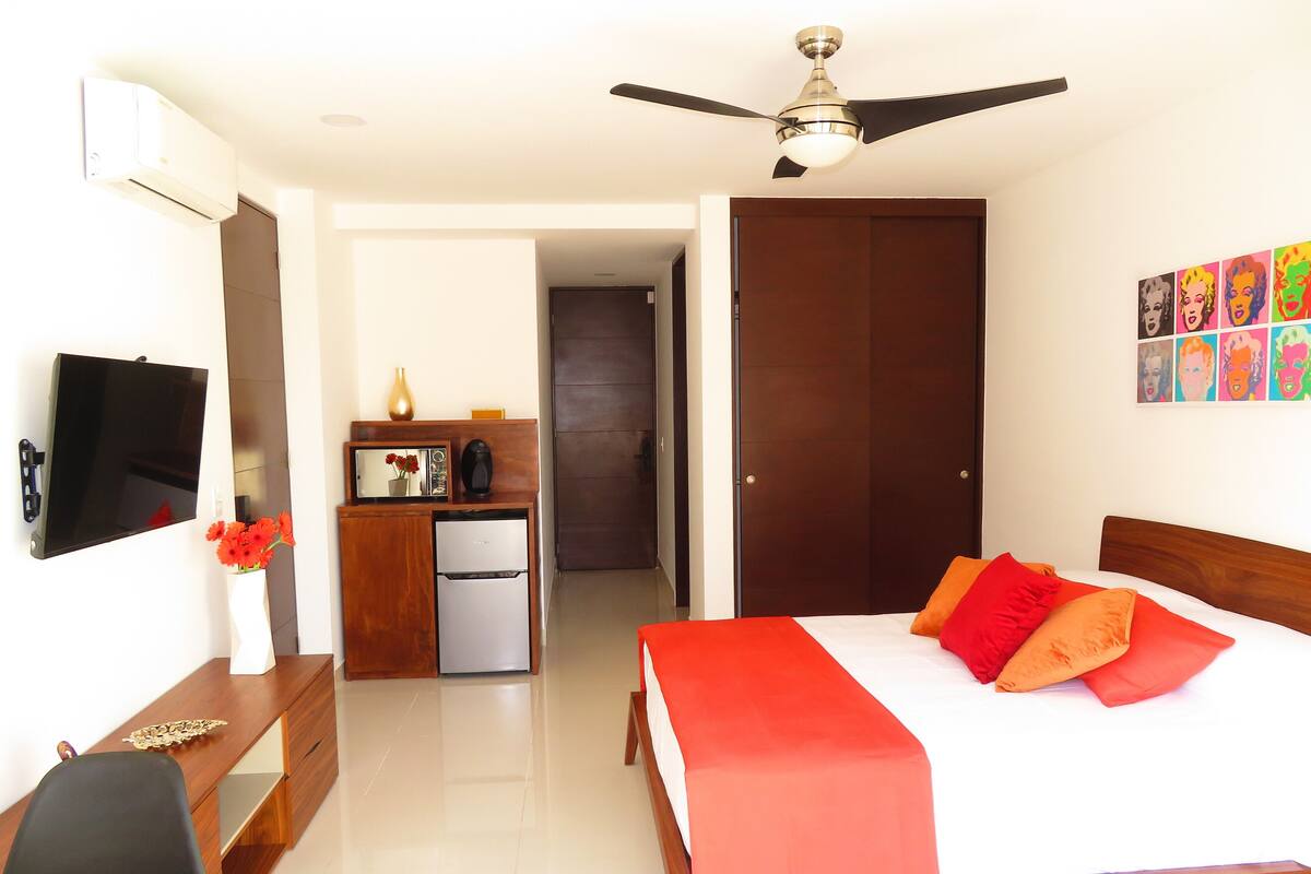 Une chambre d’hôtel comprenant un lit, un bureau, une télévision, un ventilateur de plafond, un réfrigérateur, un four à micro-ondes, un vase avec des fleurs et une porte donnant sur une autre pièce.